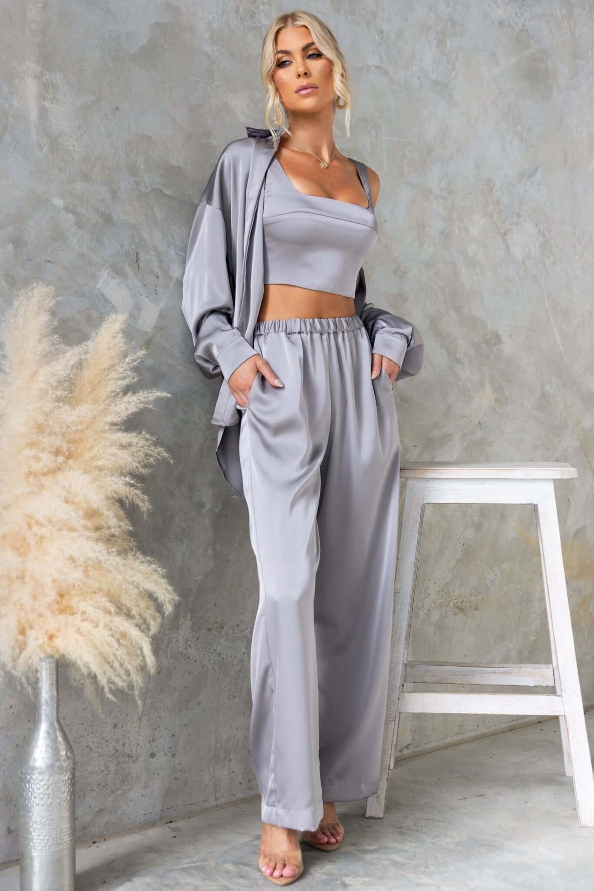 Stylish woman wearing gray satin outfit, featuring a button-up shirt and loose pants, against a textured background.