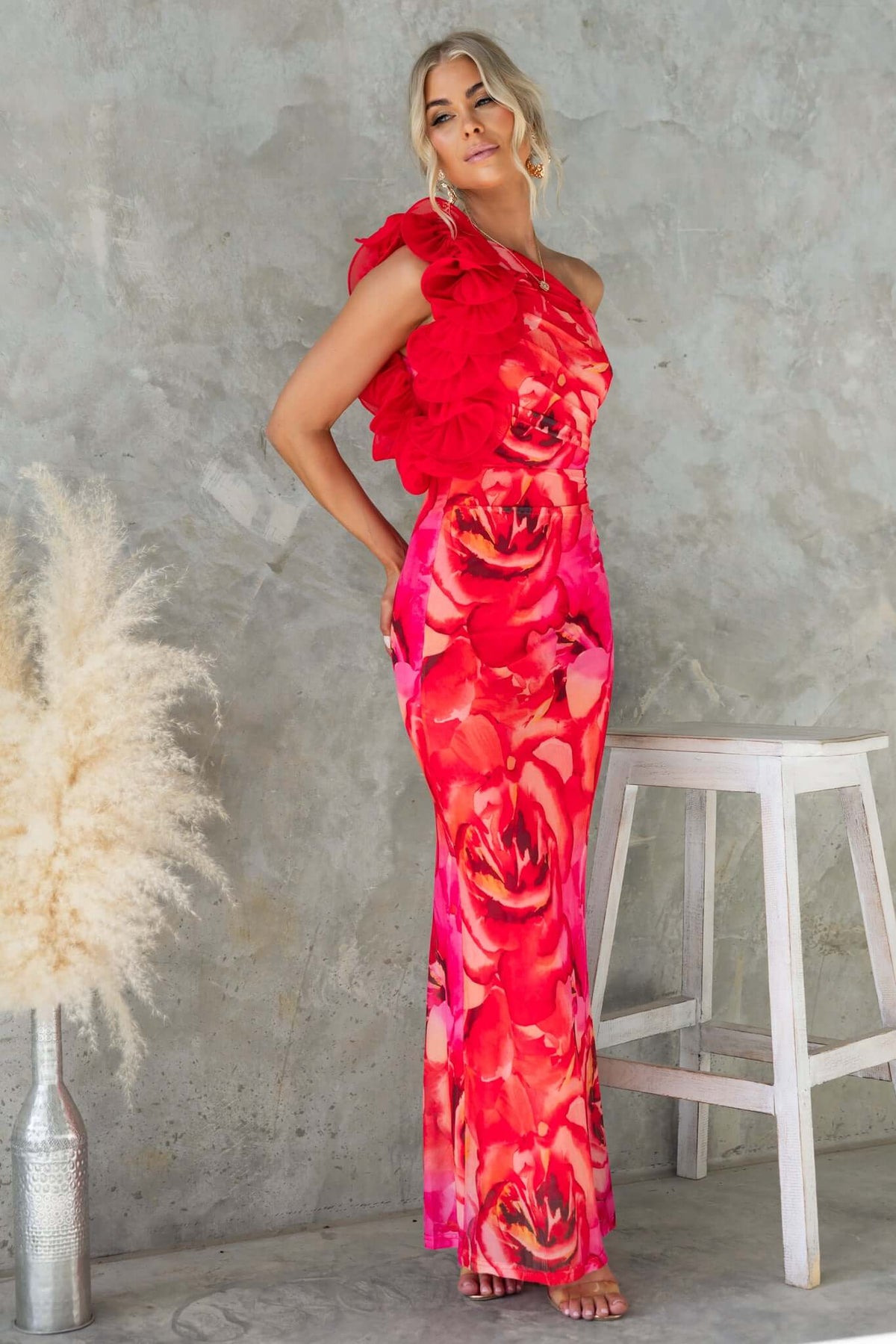 Model in vibrant red floral Meriden Maxi Dress with one-shoulder neckline and ruffle detail, styled against a textured wall.