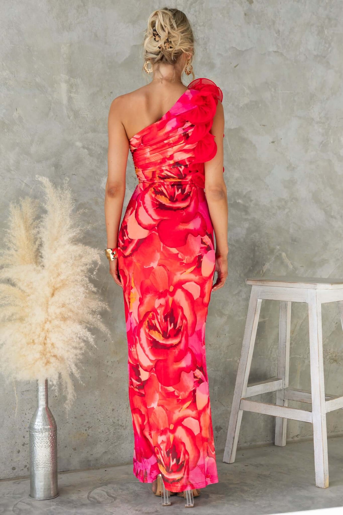 Model wearing Meriden Maxi Dress, vibrant floral design, one-shoulder neckline, standing against a textured background.