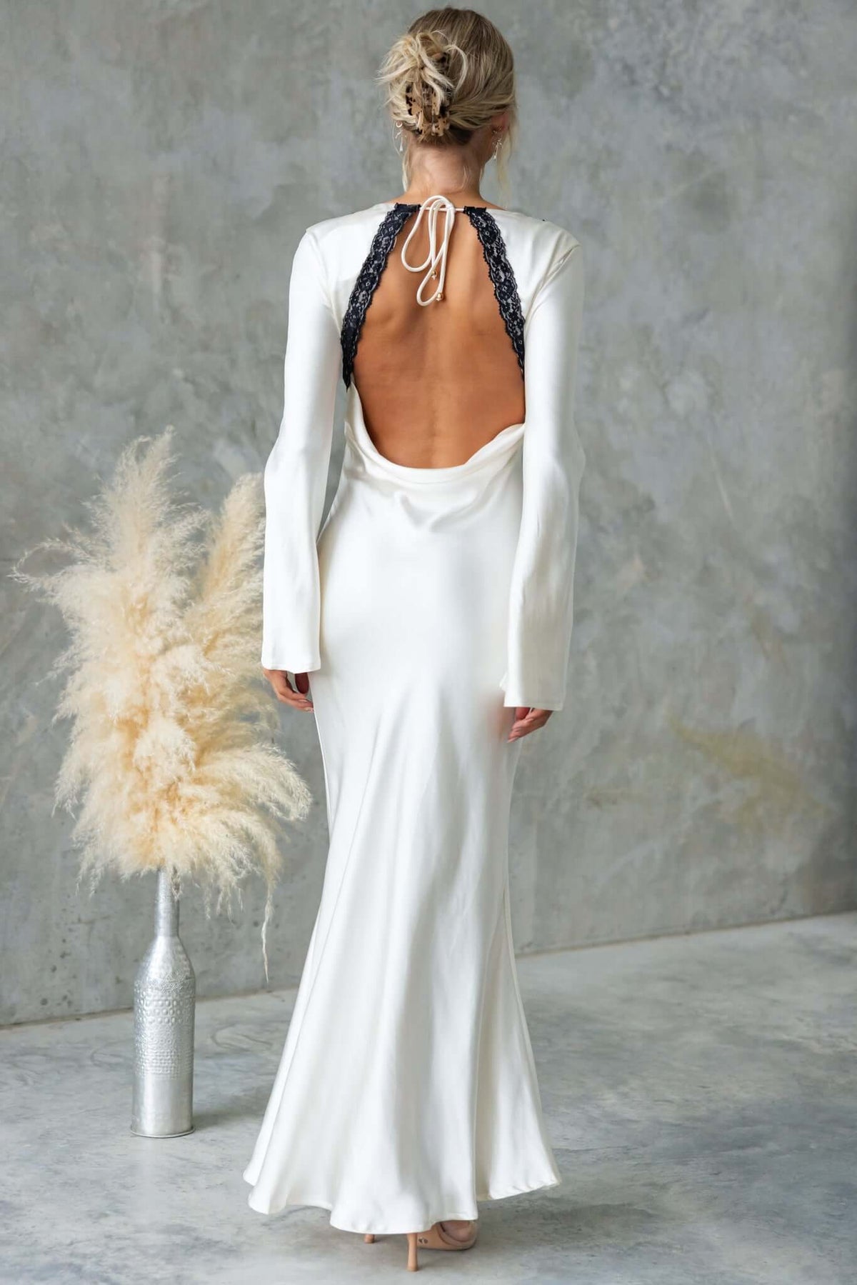 Back view of Nicholie Maxi Dress featuring lace detail, tie back, and flowing skirt, styled with beige backdrop and vase.