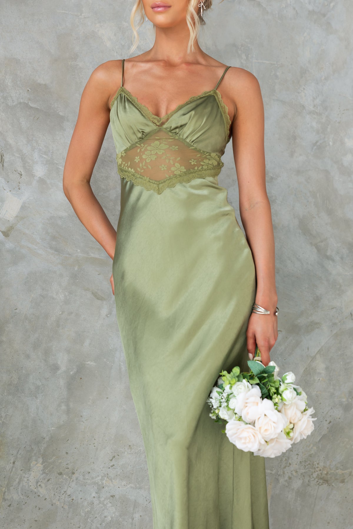 Model wearing an olive green maxi dress with lace panel and adjustable straps, holding a bouquet of white flowers.