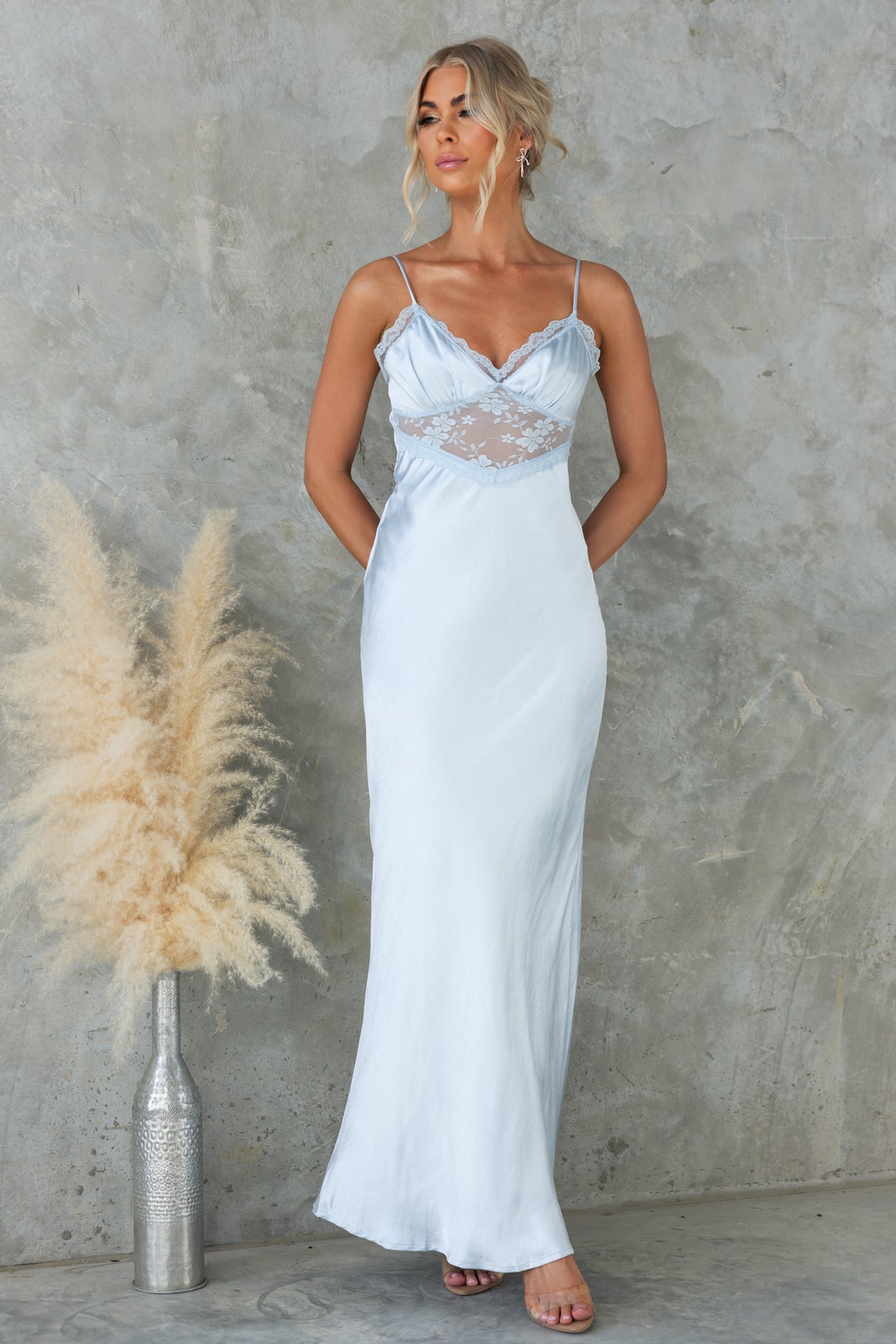 Model wearing a light blue Obsidian Maxi Dress with lace panel and adjustable straps, styled against a textured wall.