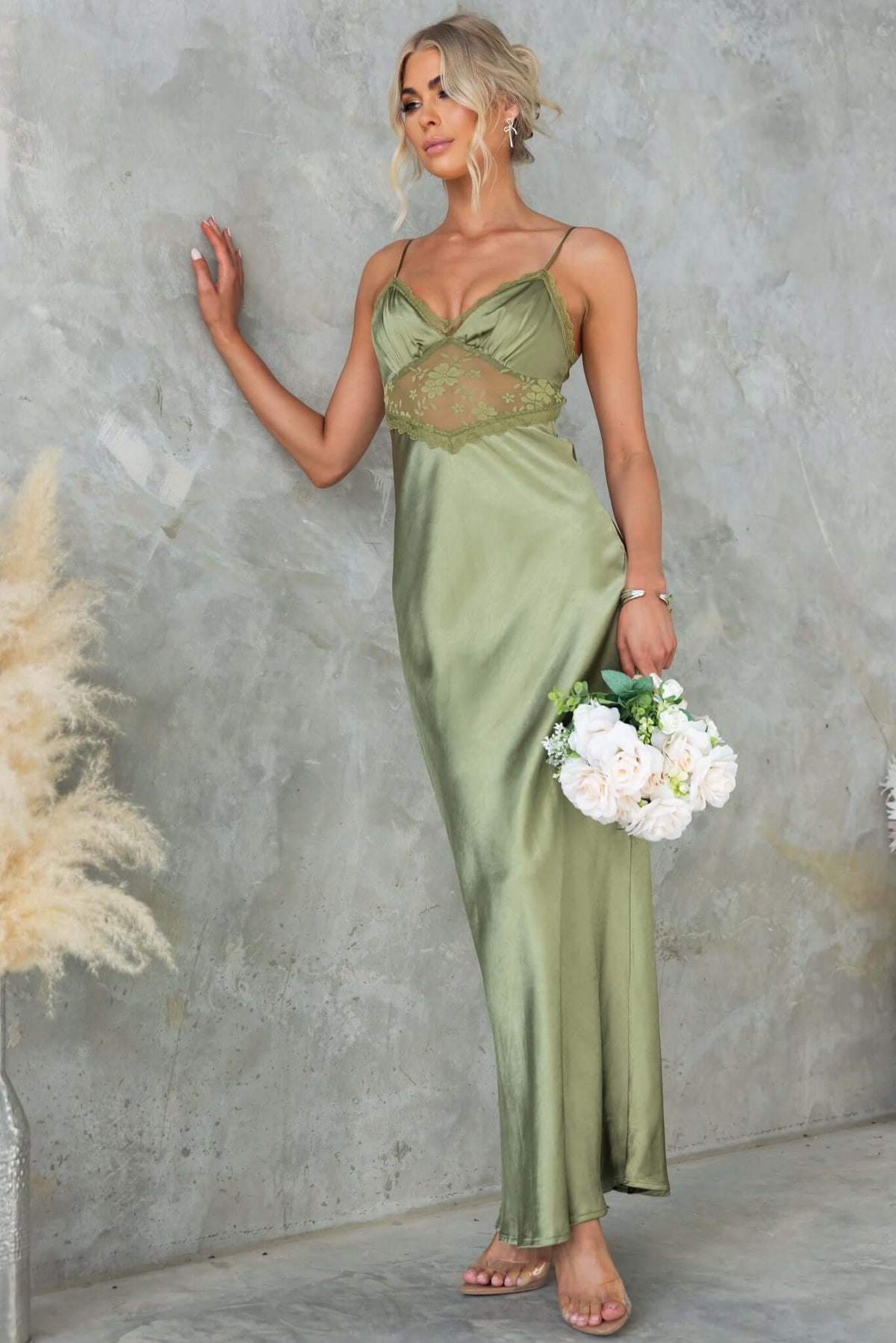 Model wearing a chic olive green maxi dress with lace panel and adjustable straps, holding a bouquet of flowers.