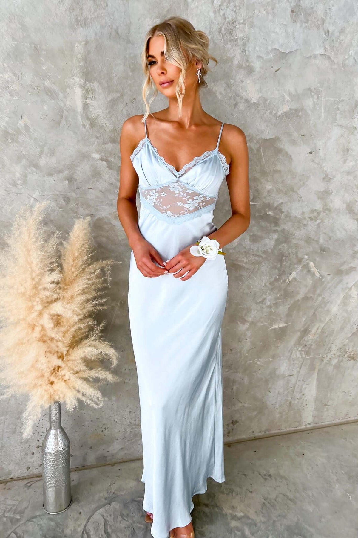 Model wearing a light blue maxi dress with lace details and adjustable straps, posed against a textured wall.