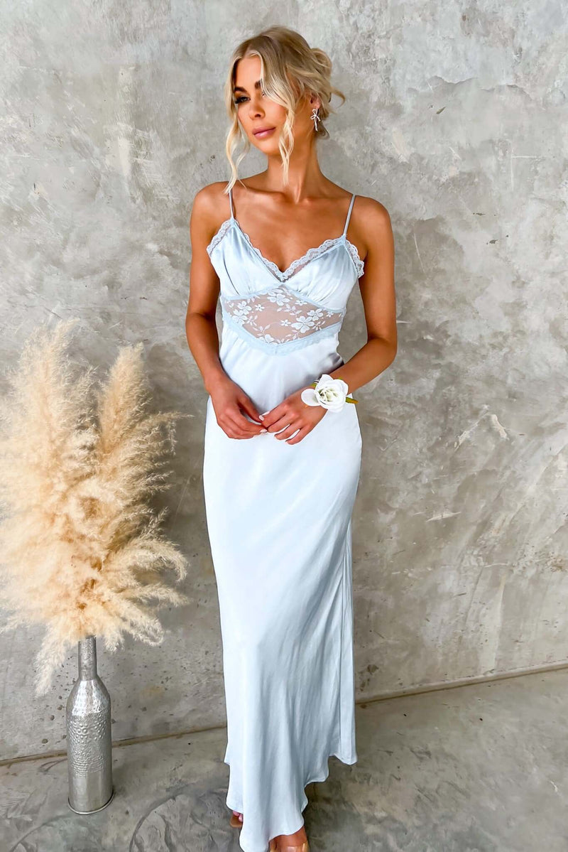Model wearing a light blue maxi dress with lace details and adjustable straps, posed against a textured wall.