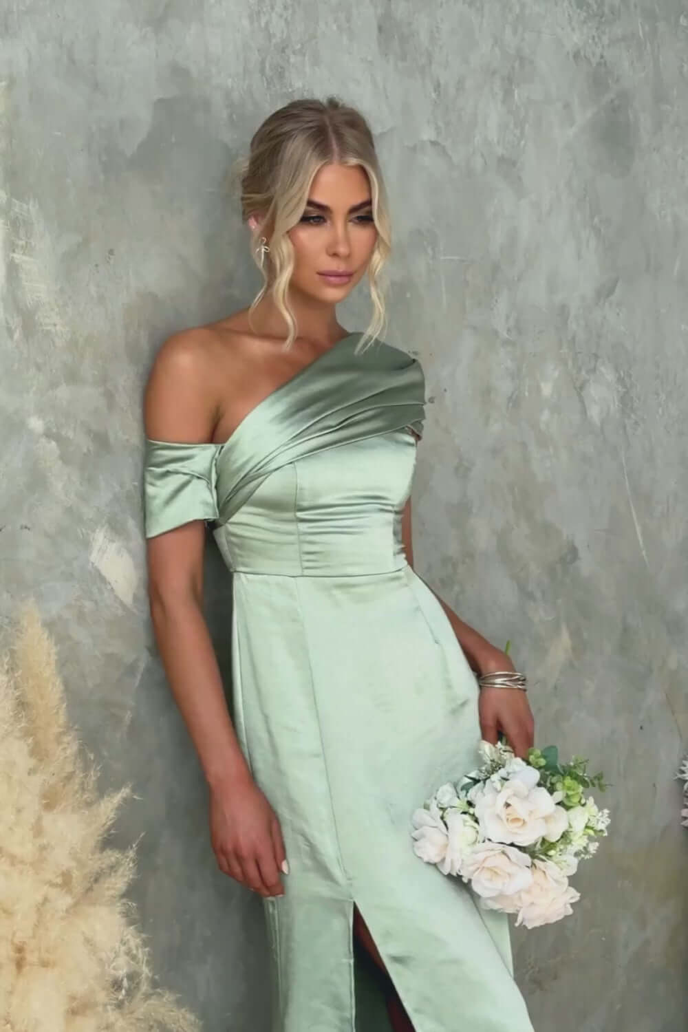 Model wearing Sonnet Maxi Dress in soft green satin with asymmetric neckline and floral bouquet.