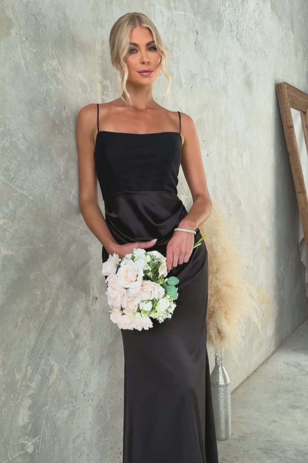 Model wearing Atheni Maxi Dress, a sleek black satin gown with adjustable straps, holding a bouquet of flowers.