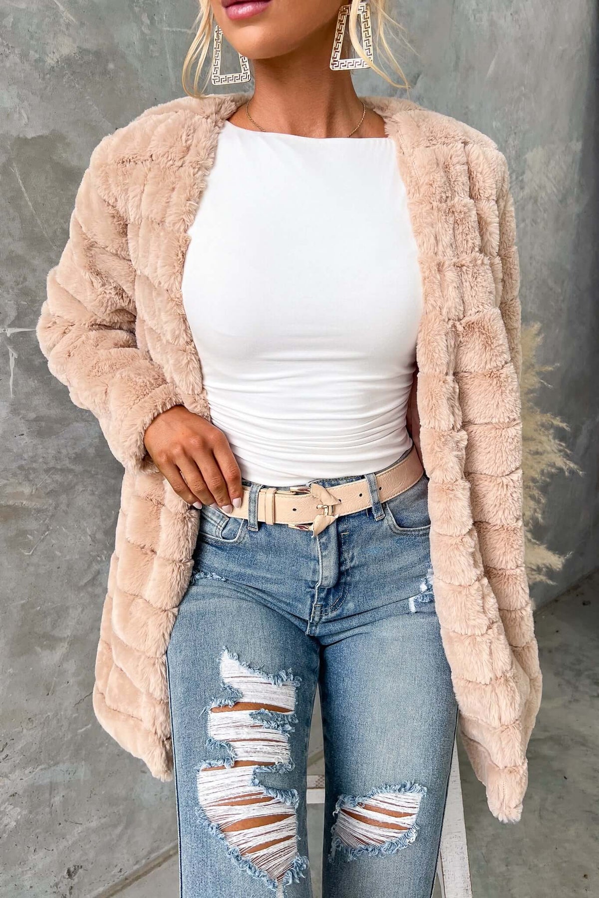 Stylish woman wearing a beige faux fur Quentin Jacket, paired with ripped jeans and a white top, showcased against a textured background.