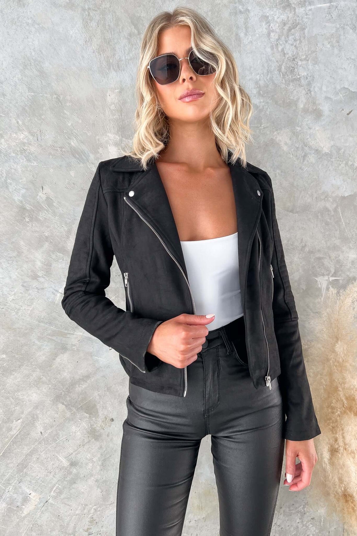 Woman in a black faux suede Remind Me Jacket with zip detailing and sunglasses, styled with high-waisted pants.