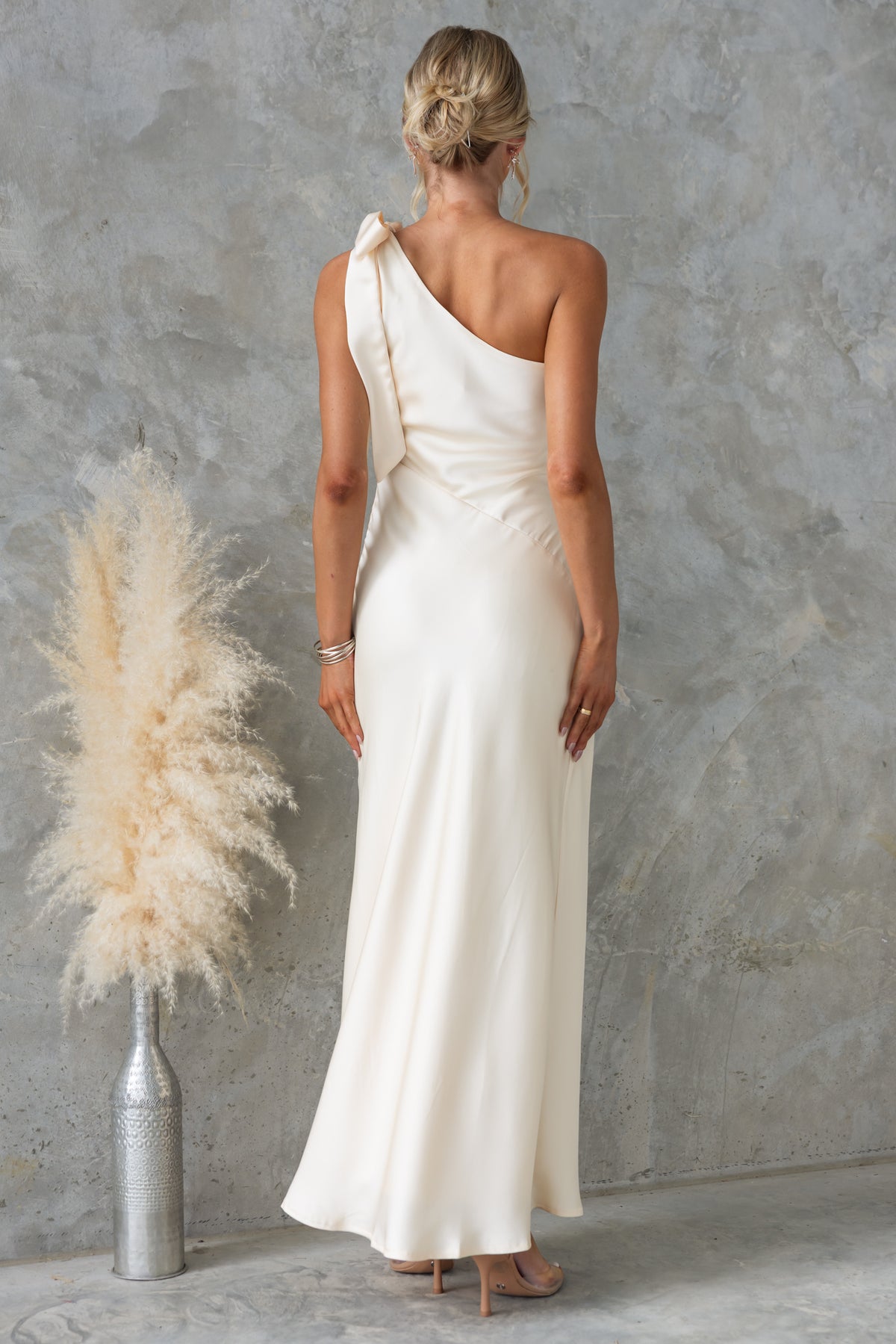 Back view of Ronnie Maxi Dress showcasing one-shoulder tie neckline and satin material against a textured backdrop.