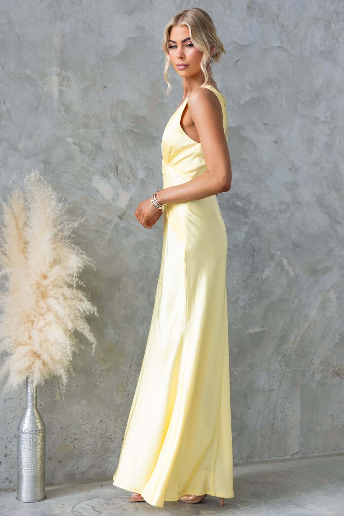 Model wearing the Uvira Maxi Dress, a satin yellow dress with a V-neckline and side zip, set against a textured backdrop.