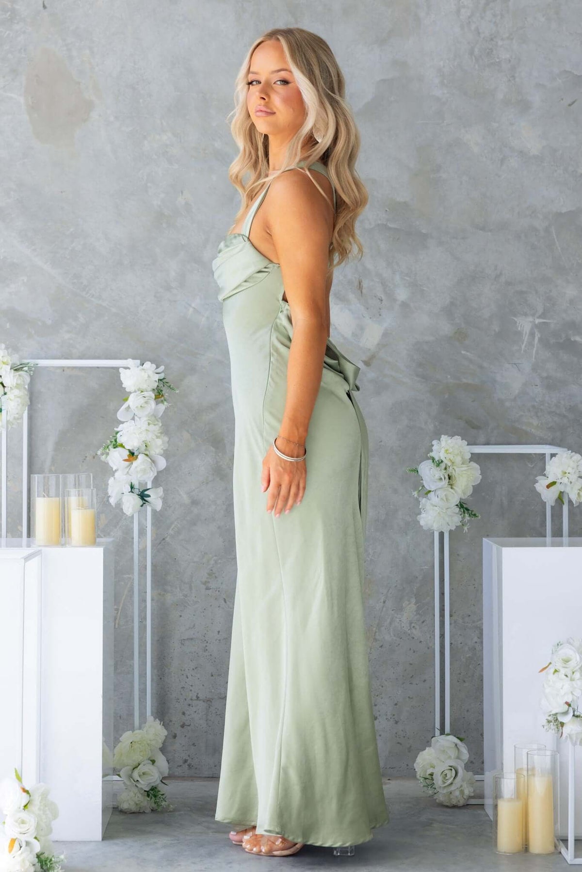 Megan Maxi Dress in soft green with adjustable tie straps, showcasing a sleek silhouette on a model against a textured background.