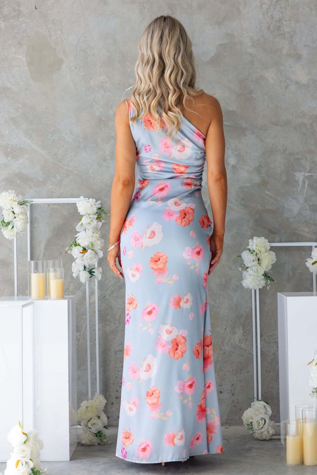 Back view of a floral print jadeite maxi dress with a one-shoulder neckline against a neutral wall.