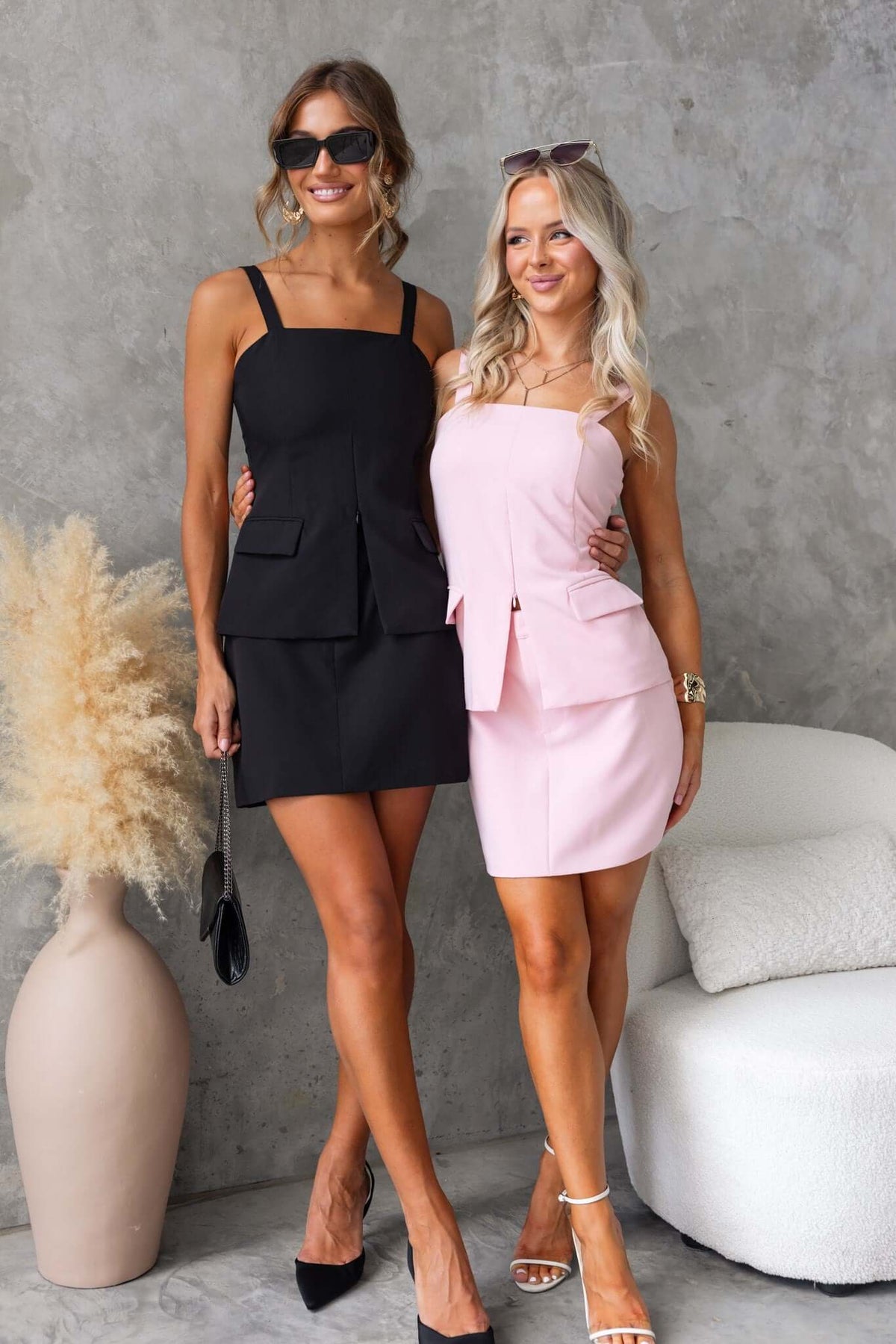 Two women posing in stylish mid-high waist skirts, one in black and the other in pink, both with faux pockets.