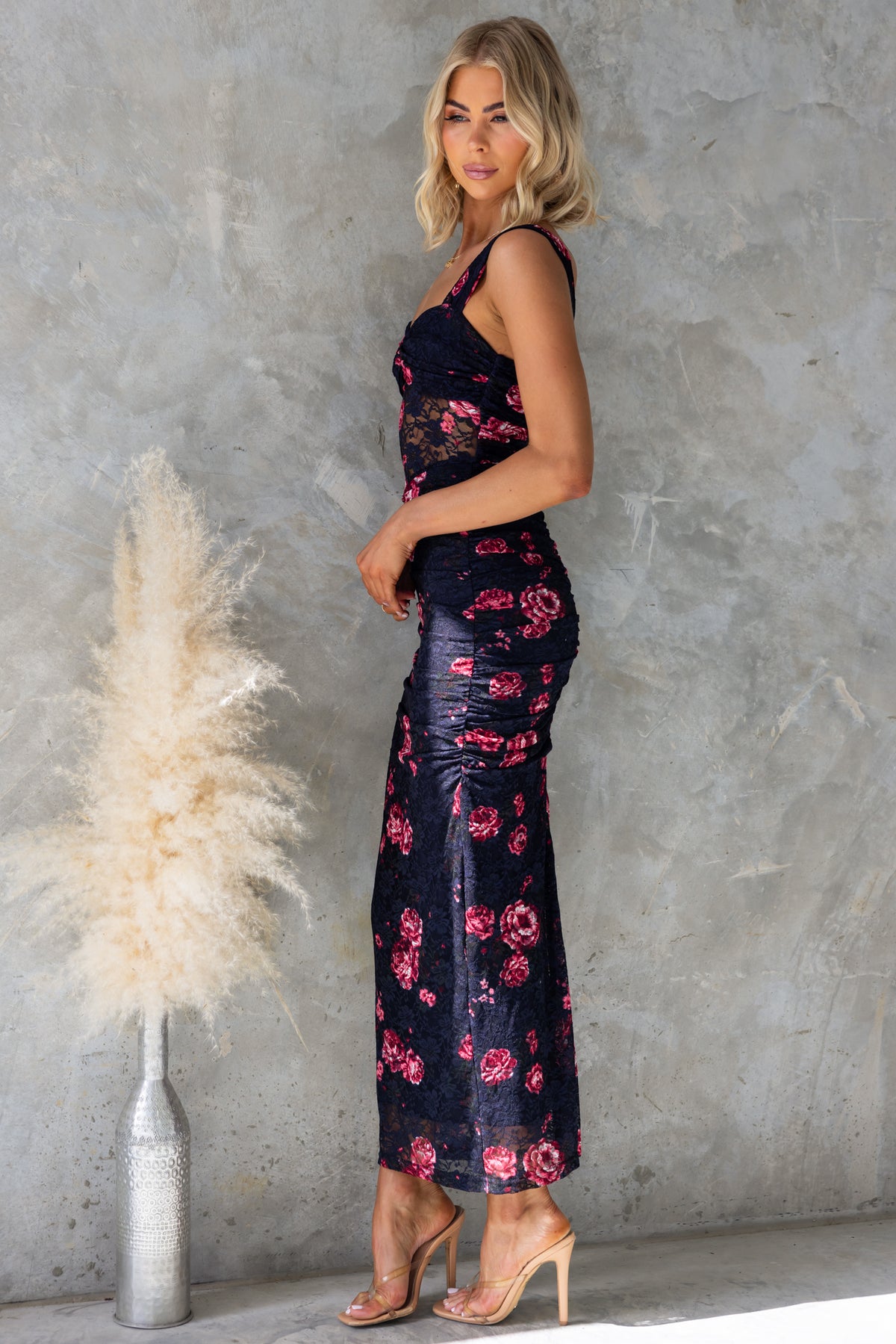 Model showcasing Xavina Maxi Dress featuring floral design, fitted silhouette, and subtle ruching against a textured background.