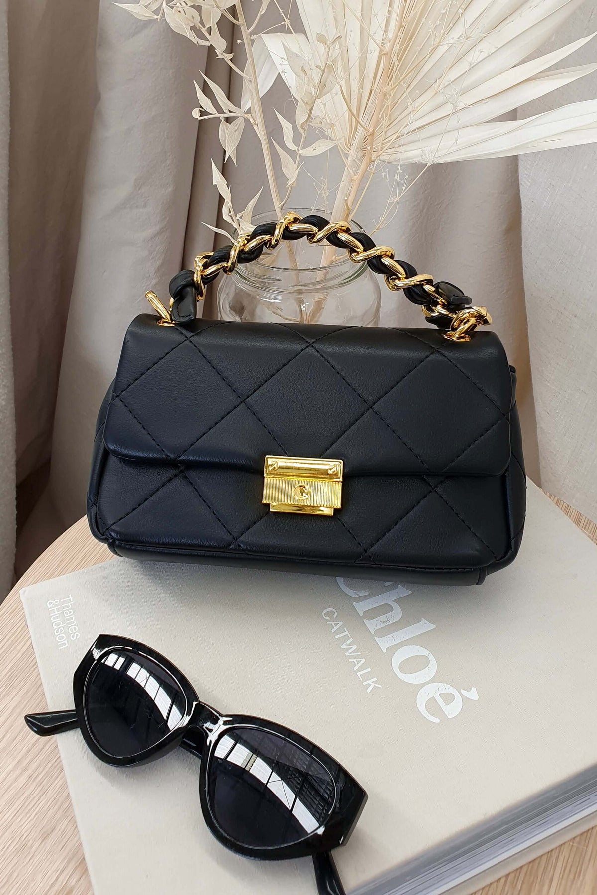 Stylish black vegan leather Ari Bag with gold chain handle and clasp, placed next to trendy sunglasses on a book.