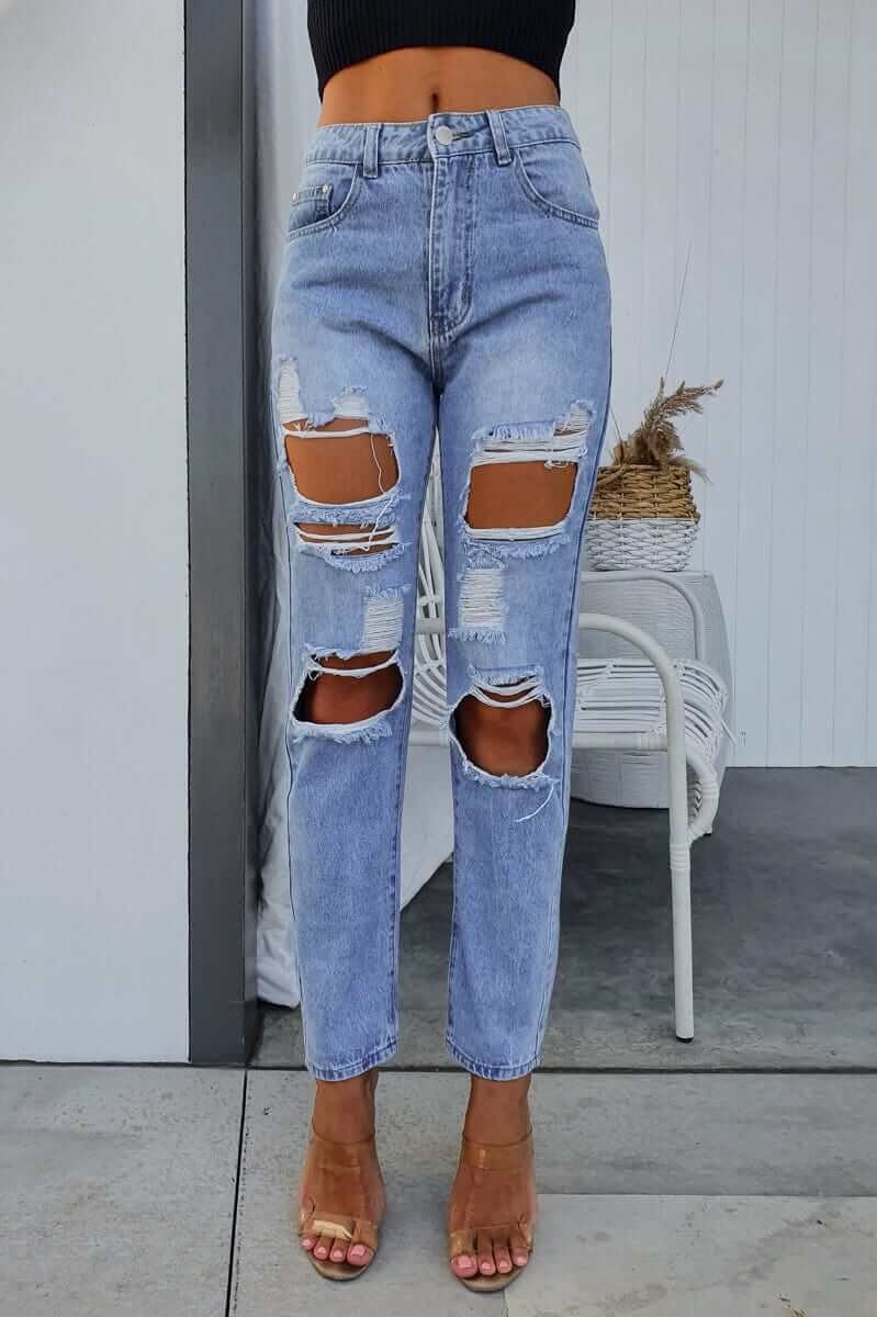 Light blue distressed mom jeans with ripped details, styled with a black top and sandals in a casual setting.