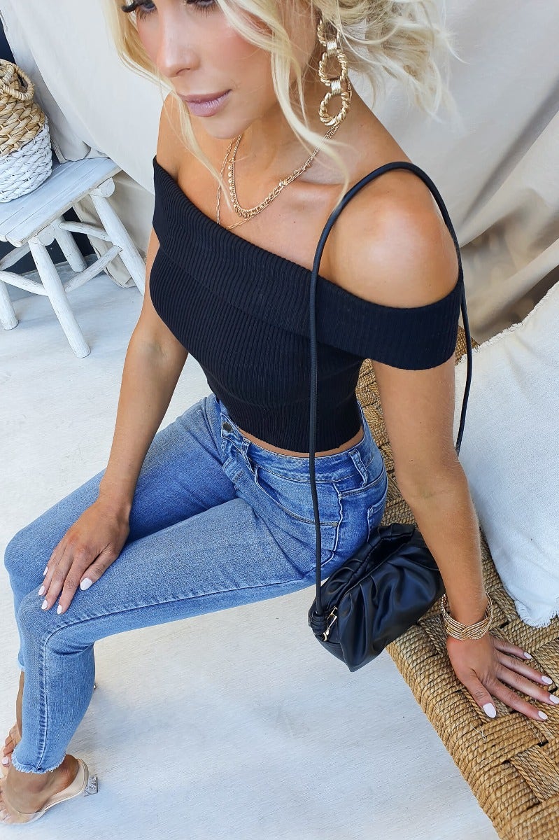 Model wearing a black ribbed knit off-shoulder top paired with blue jeans and a black bag, showcasing a casual chic look.