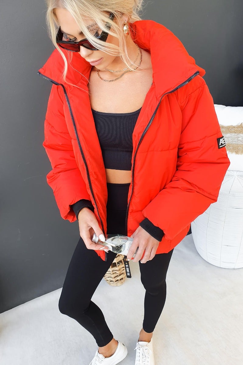 Model wearing Delila Puffer Jacket in red, featuring a high neckline and removable hood, paired with black leggings.