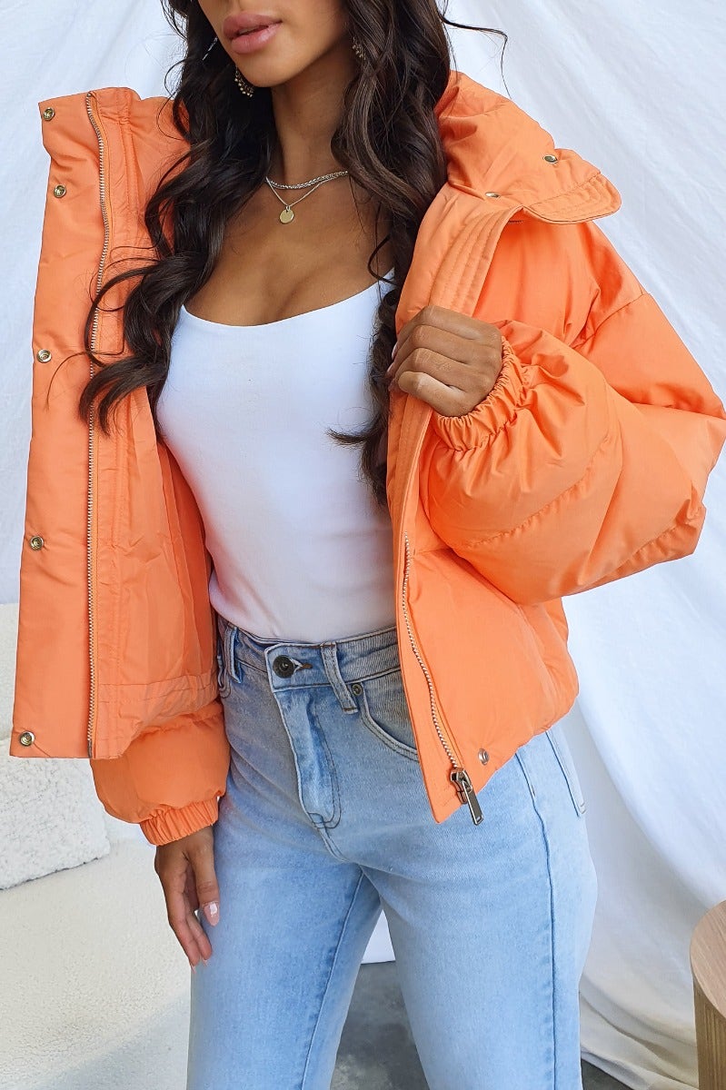 Model wearing an orange Avril Puffer Jacket with high neckline, front closure, and elastic cuffs, paired with jeans.