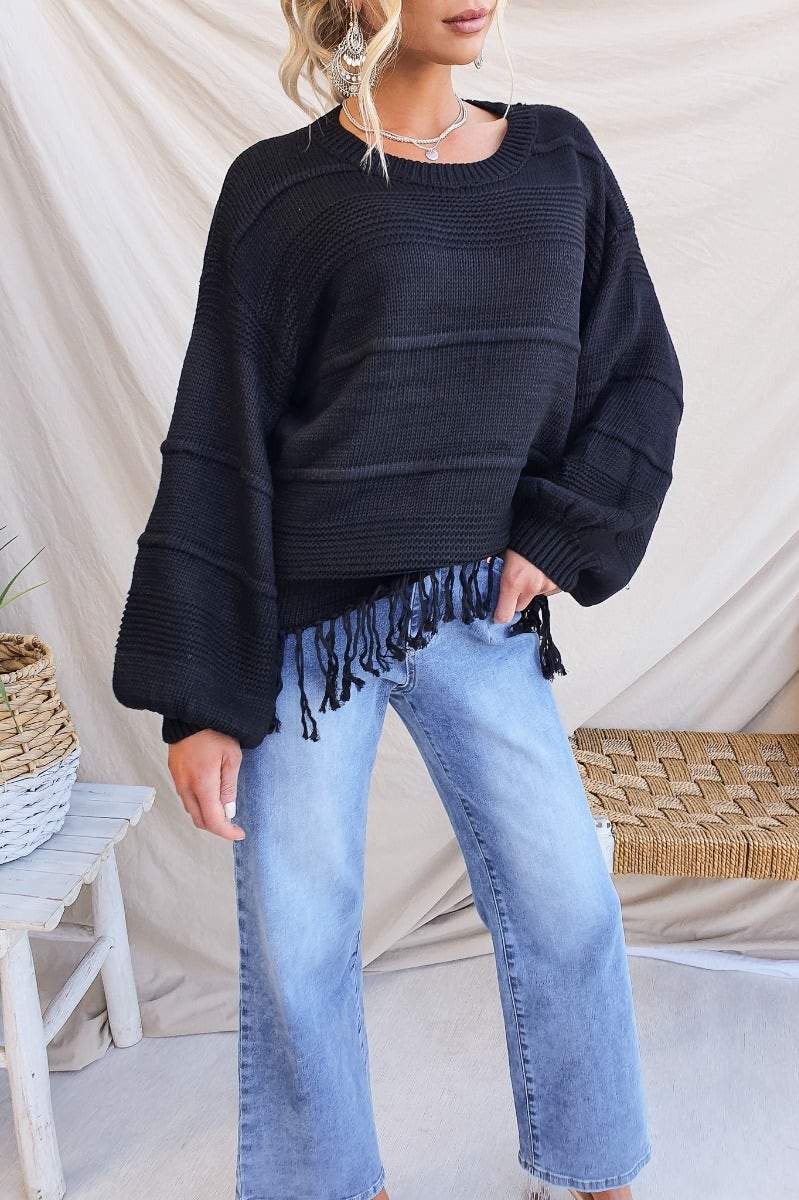 Model wearing a black Adela Knit jumper with a round neckline and tassle detail, paired with light blue jeans.