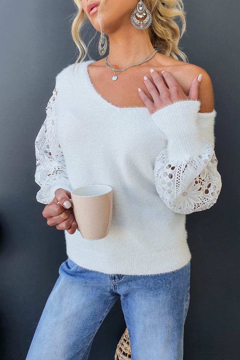 Woman wearing white Indya jumper with lace sleeves, V-neckline, holding a mug, styled with casual jeans and accessories.