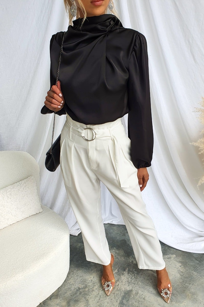 Black satin Xali top with high neckline and fitted sleeves paired with white trousers in a stylish indoor setting.