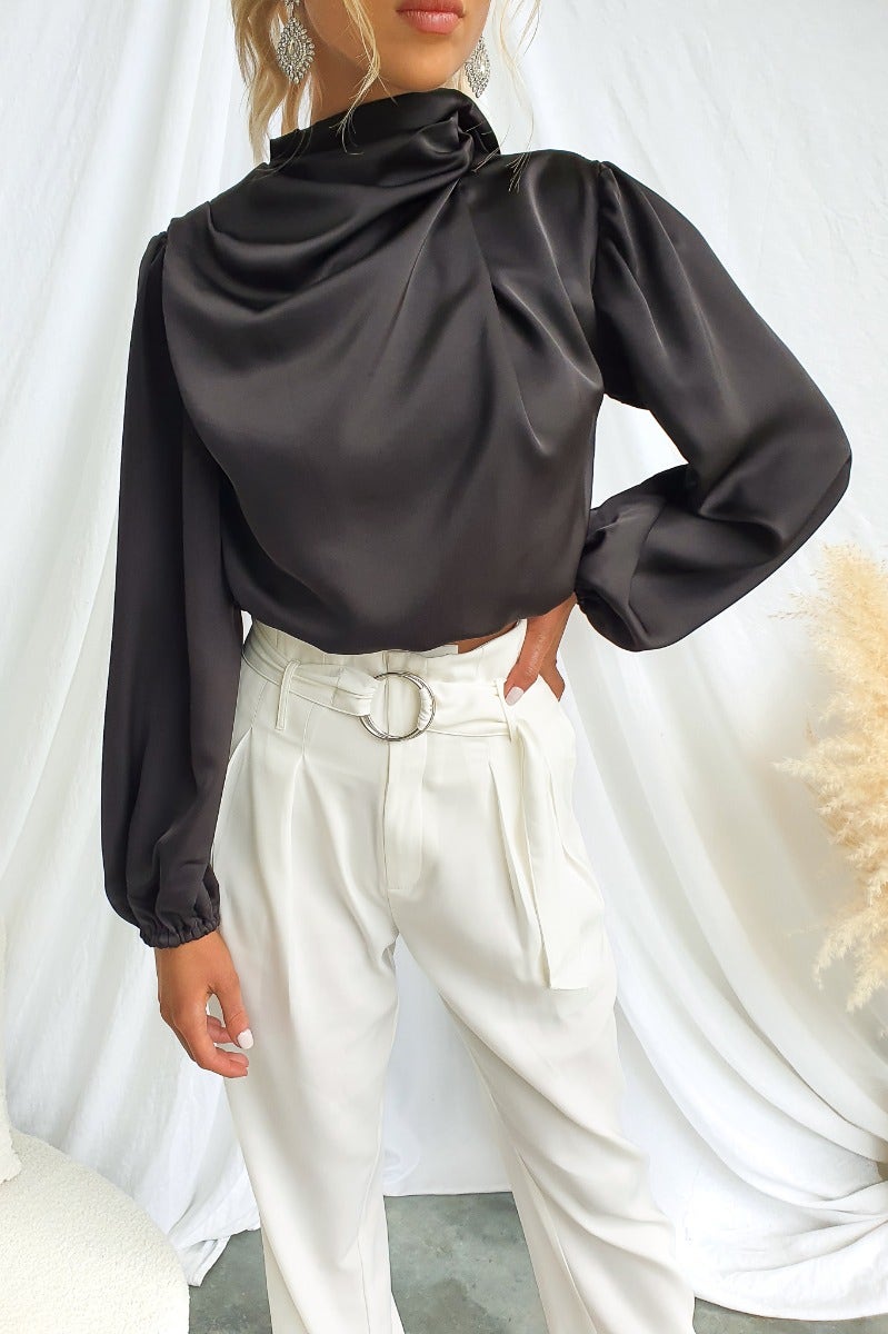 Black satin Xali Top featuring a high neckline, long sleeves, and styled with white trousers.