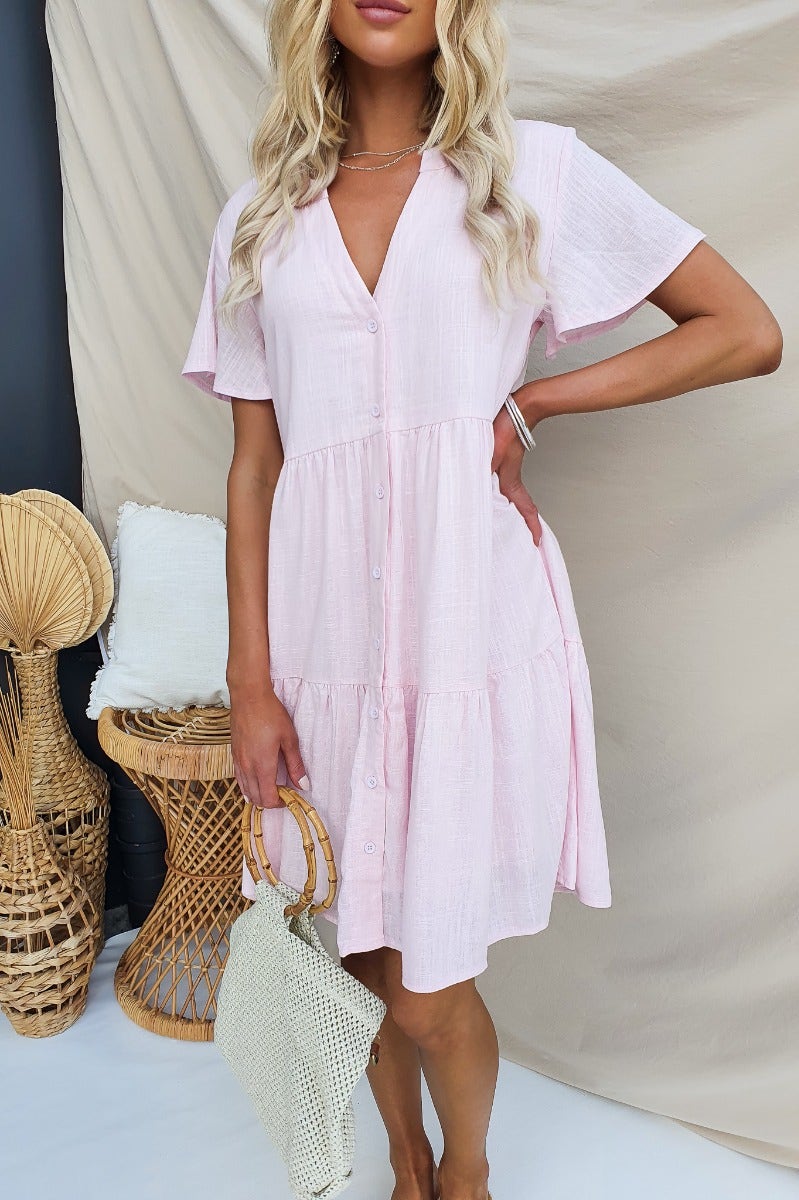 Model wearing a pink linen blend Arla Dress with V-neckline and button-up front, styled with a woven bag.