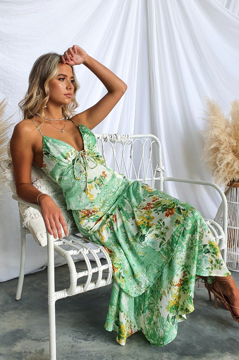Model wearing a floral print Wildest Dreams maxi dress with adjustable straps, sitting on a chair near decorative plants.
