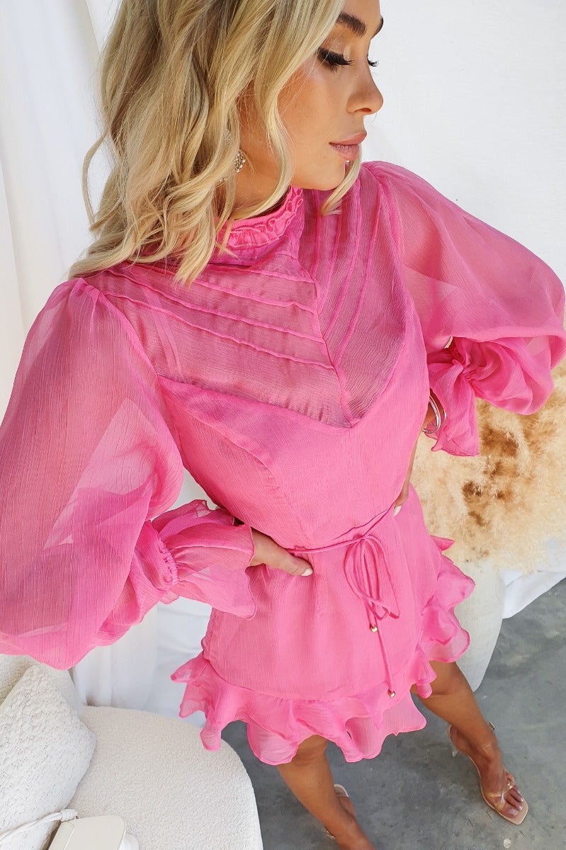 Model wearing a pink chiffon Encore Dress with puff sleeves and ruffled hem, showcasing a stylish high neckline and tie detail.