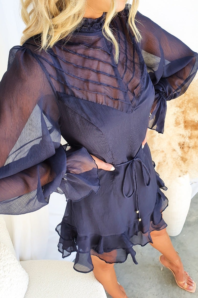 Navy chiffon Encore dress with high neckline, elastic sleeves, and removable tie, styled with ruffled hem.