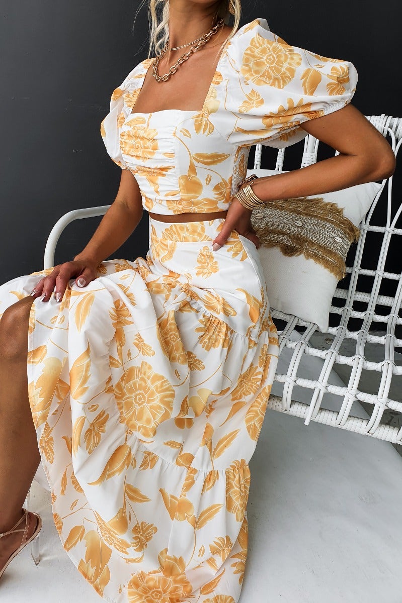 Model wearing Somebody To Love floral print set with puffy sleeves and a maxi skirt, seated on a stylish white chair.