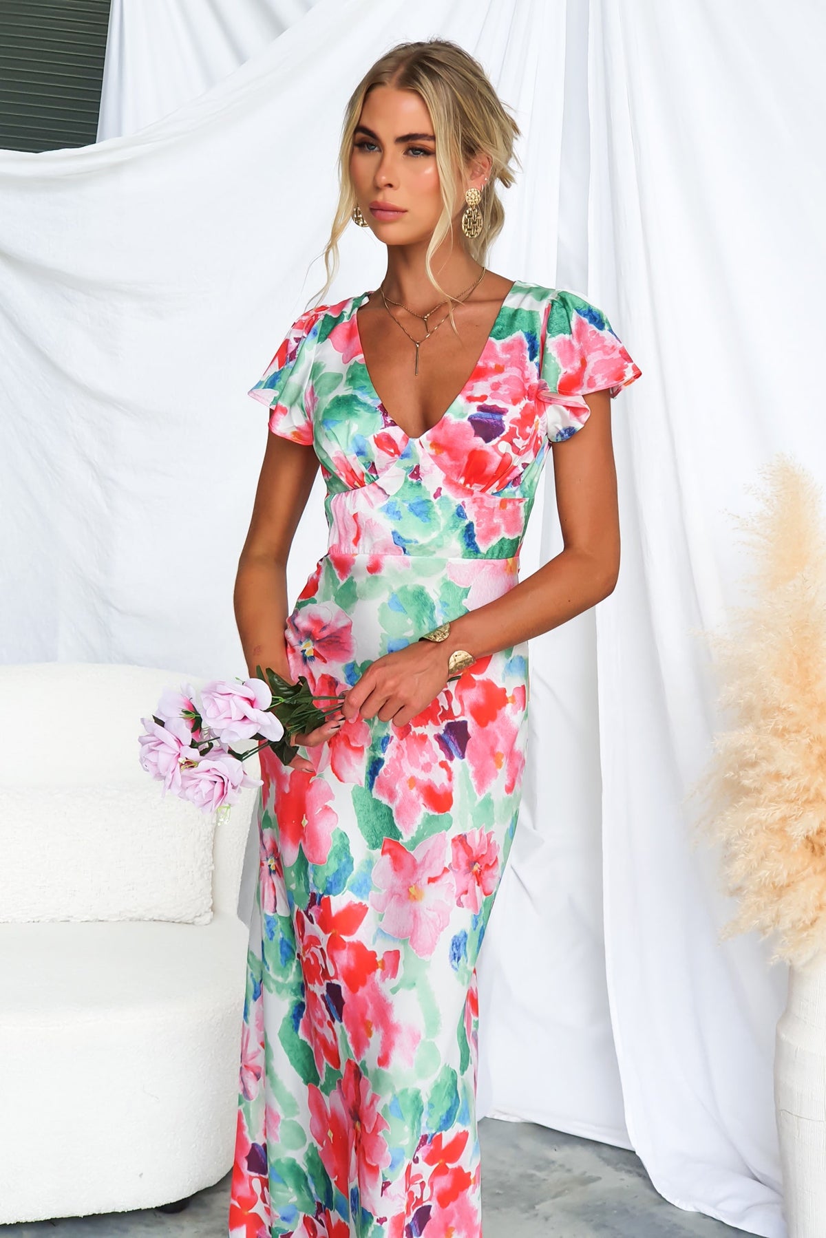 Model wearing a floral satin Belmont Maxi Dress with cap sleeves, standing elegantly and holding flowers.