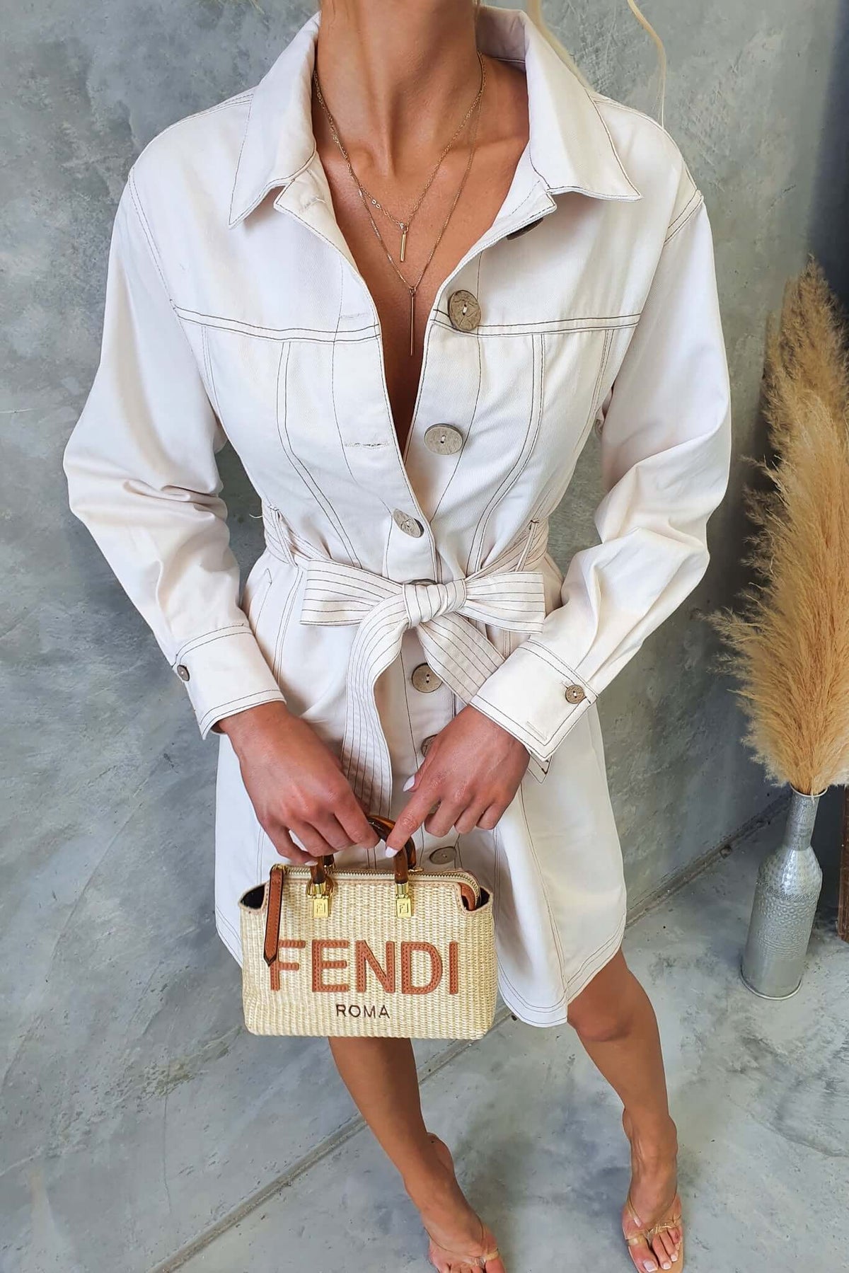 Model wearing a white denim dress with buttons and a waist belt, holding a designer bag, in a stylish indoor setting.
