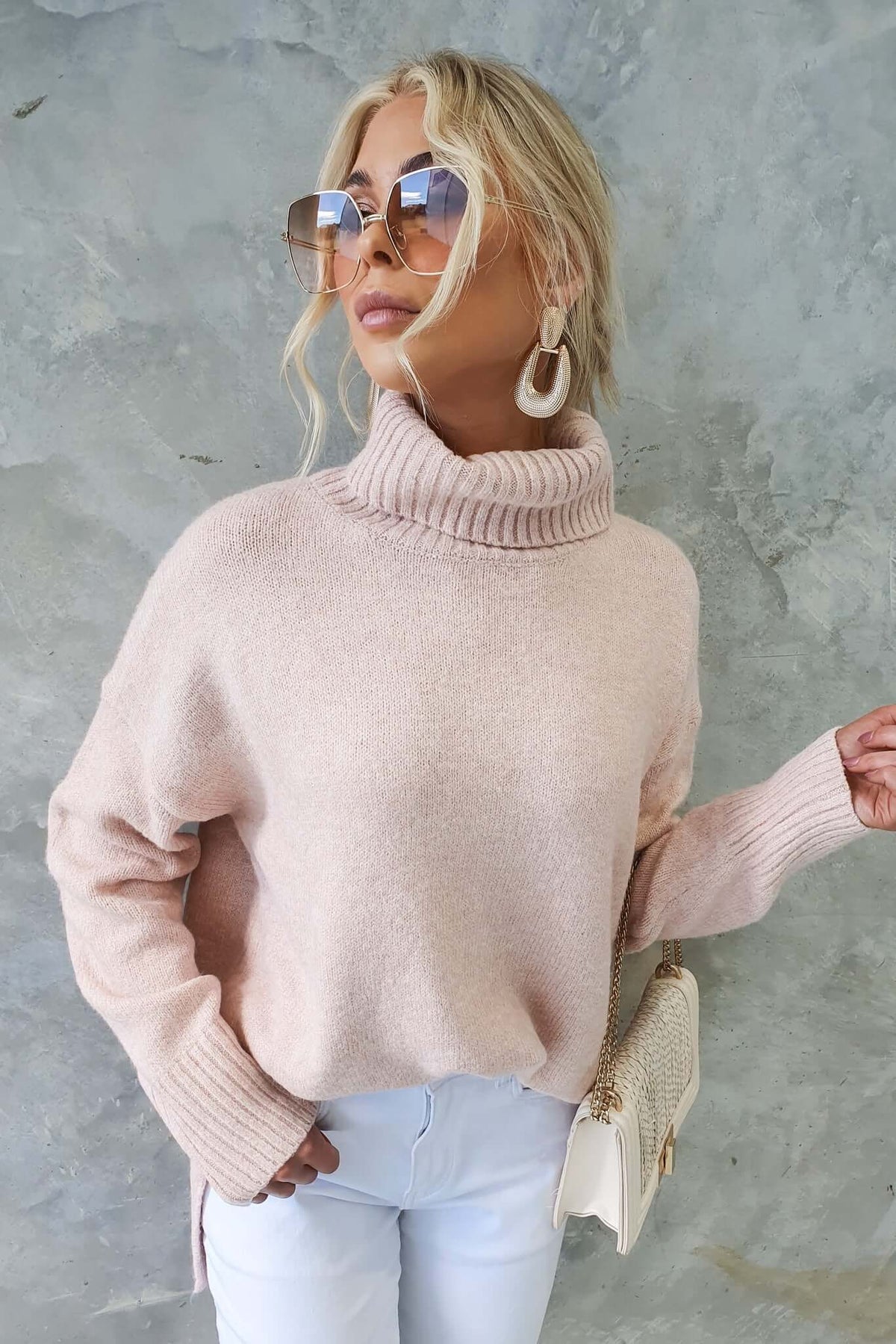 Model wearing a light pink ribbed knit turtle neck jumper with relaxed fit and small side splits, accessorized with large sunglasses.