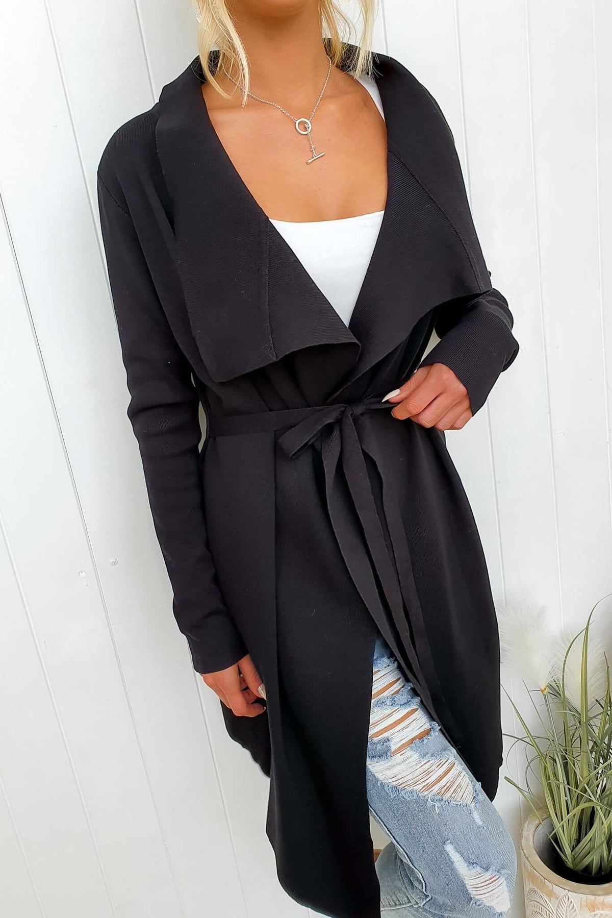 Acaia Cardigan in black ribbed knit, featuring a collar, lapel, pockets, and attached belt, worn over ripped jeans.