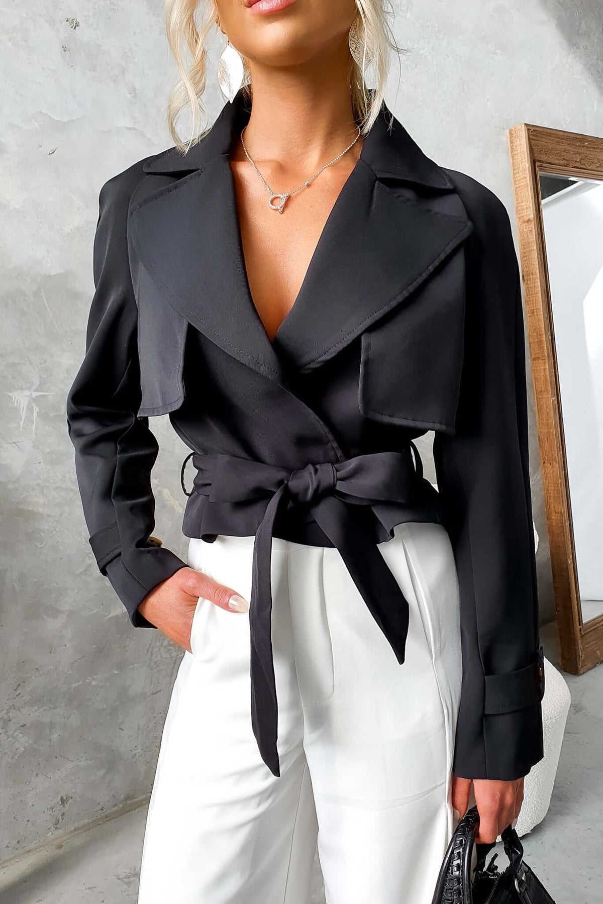 Black trench coat with a tie belt, collar, and lapel, styled with white trousers and a chic, modern look.