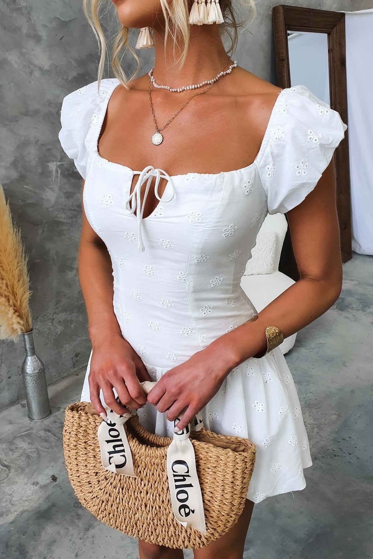 Model wearing white Fairfield Dress with puff sleeves, tie at bust, and holding a straw bag.