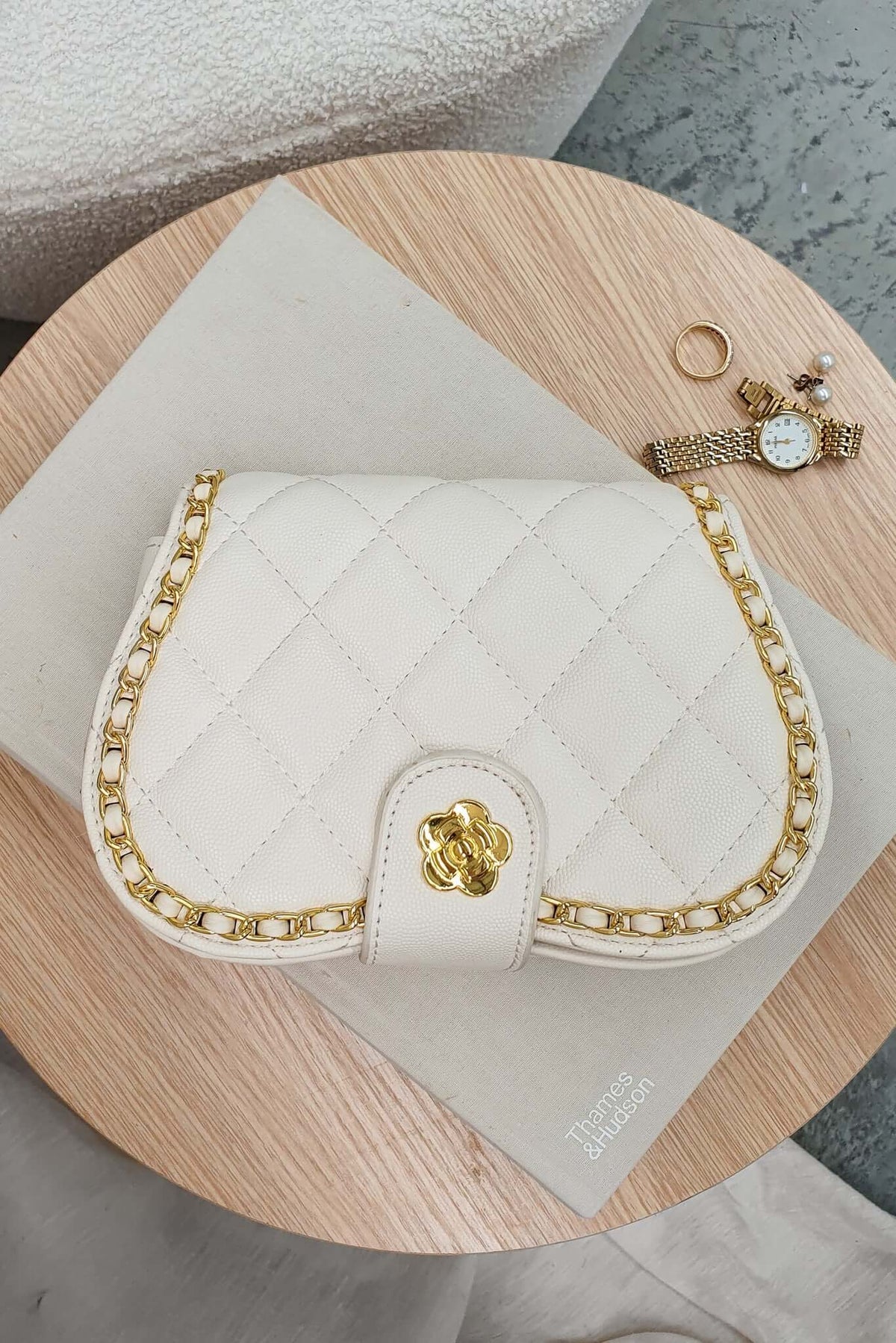 Chic white quilted handbag with gold chain, placed on a wooden surface beside a ring and watch.