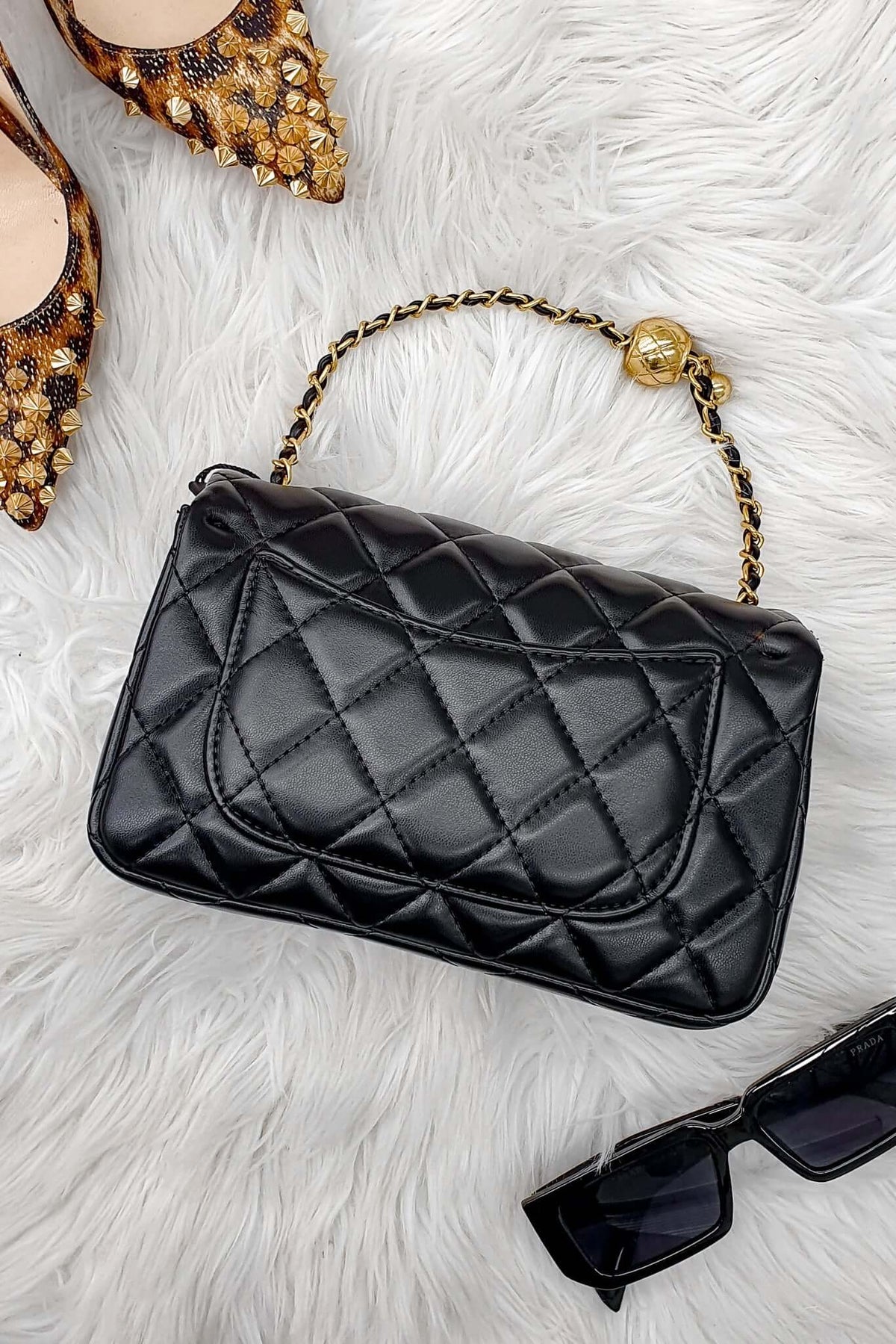 Chic black quilted Chelsea bag with gold hardware and stylish chain strap on a soft white fur background.