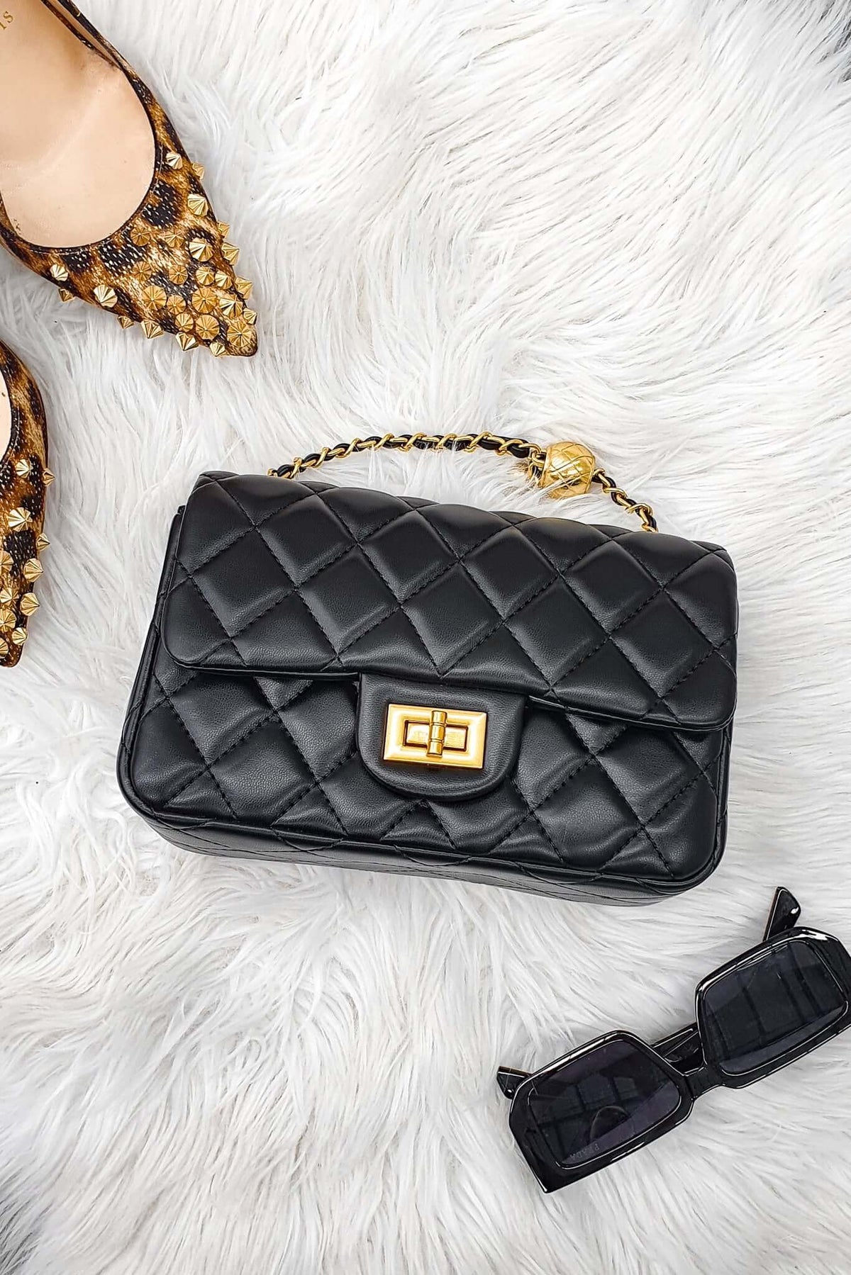 Chic black quilted Chelsea bag with gold hardware on faux fur, paired with stylish shoes and sunglasses.