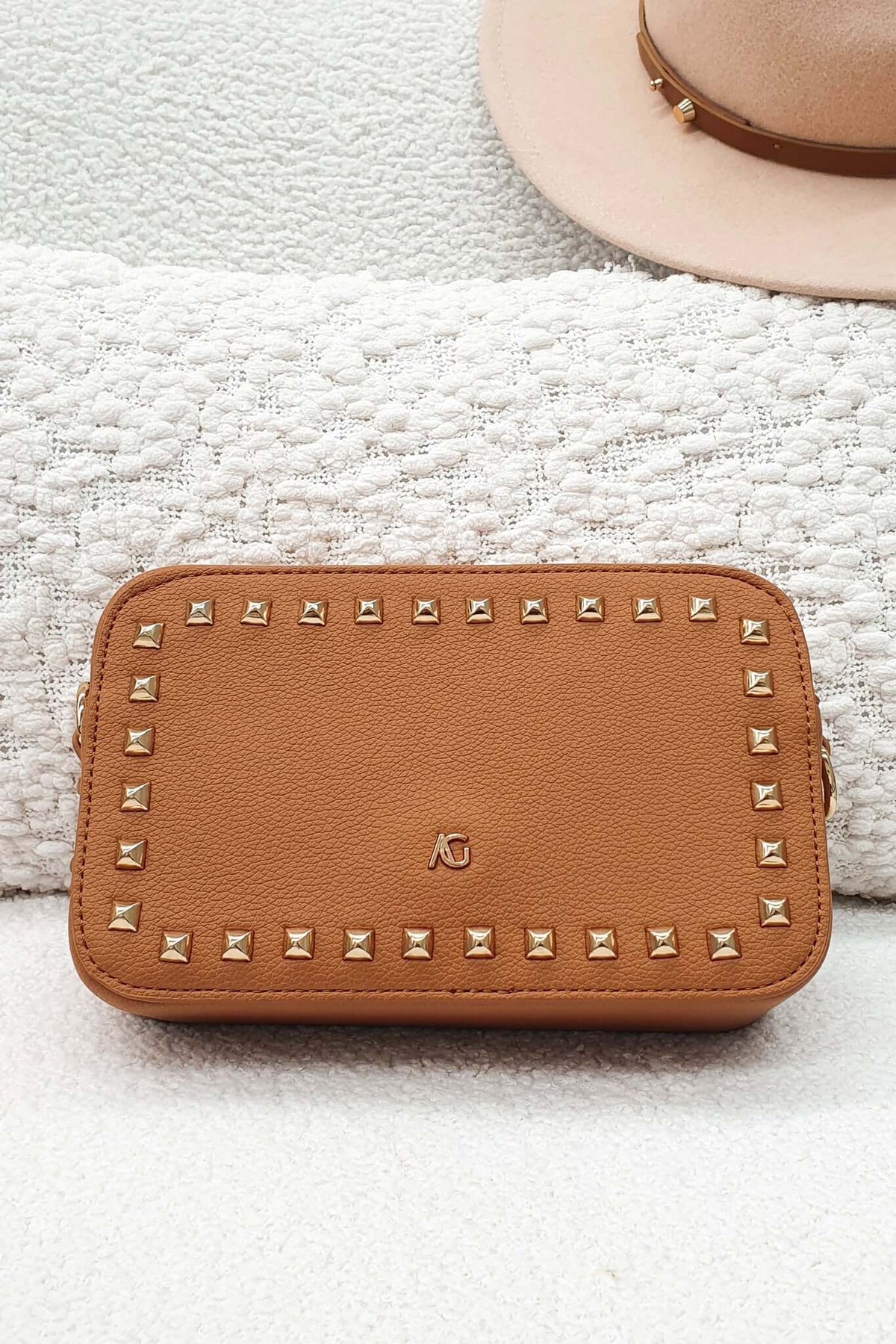 Veronica Crossbody Bag in tan leather with metallic studs, stylishly placed on a textured background.
