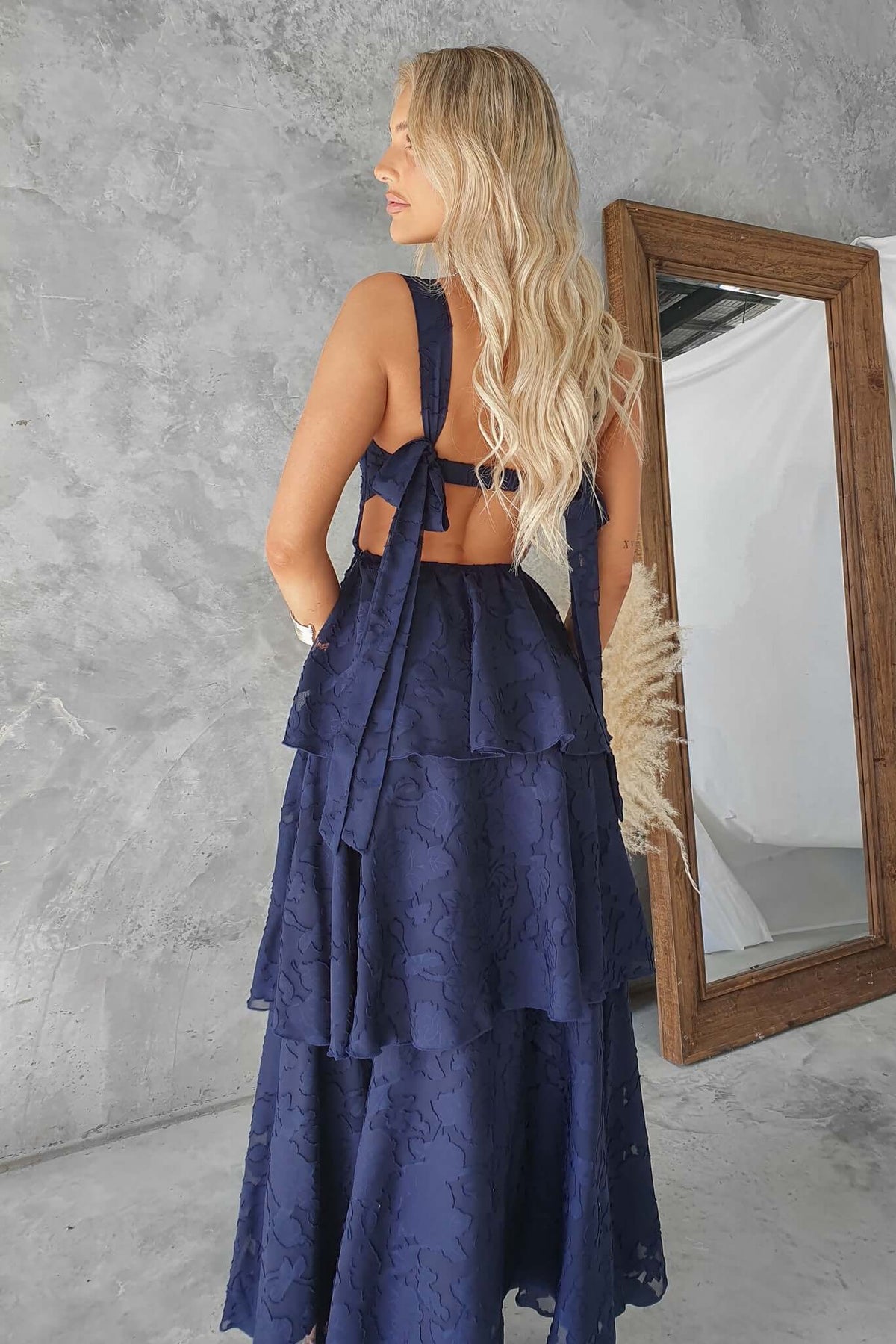 Model wearing a navy Rivoli Maxi Dress with tie straps and a ruffle skirt, standing near a mirror.