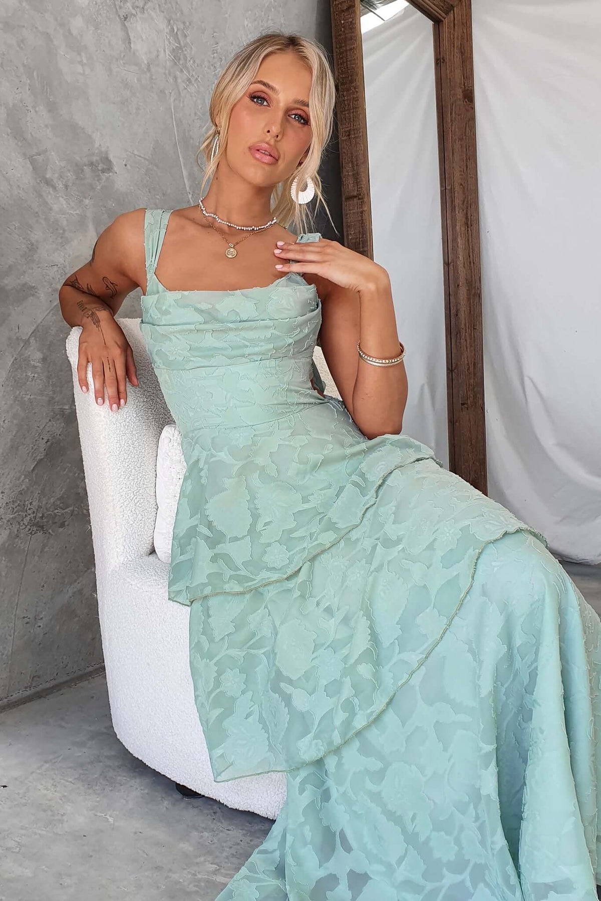 Model wearing a light blue Rivoli Maxi Dress with ruffle skirt, cowl neckline, and tie straps, sitting elegantly.
