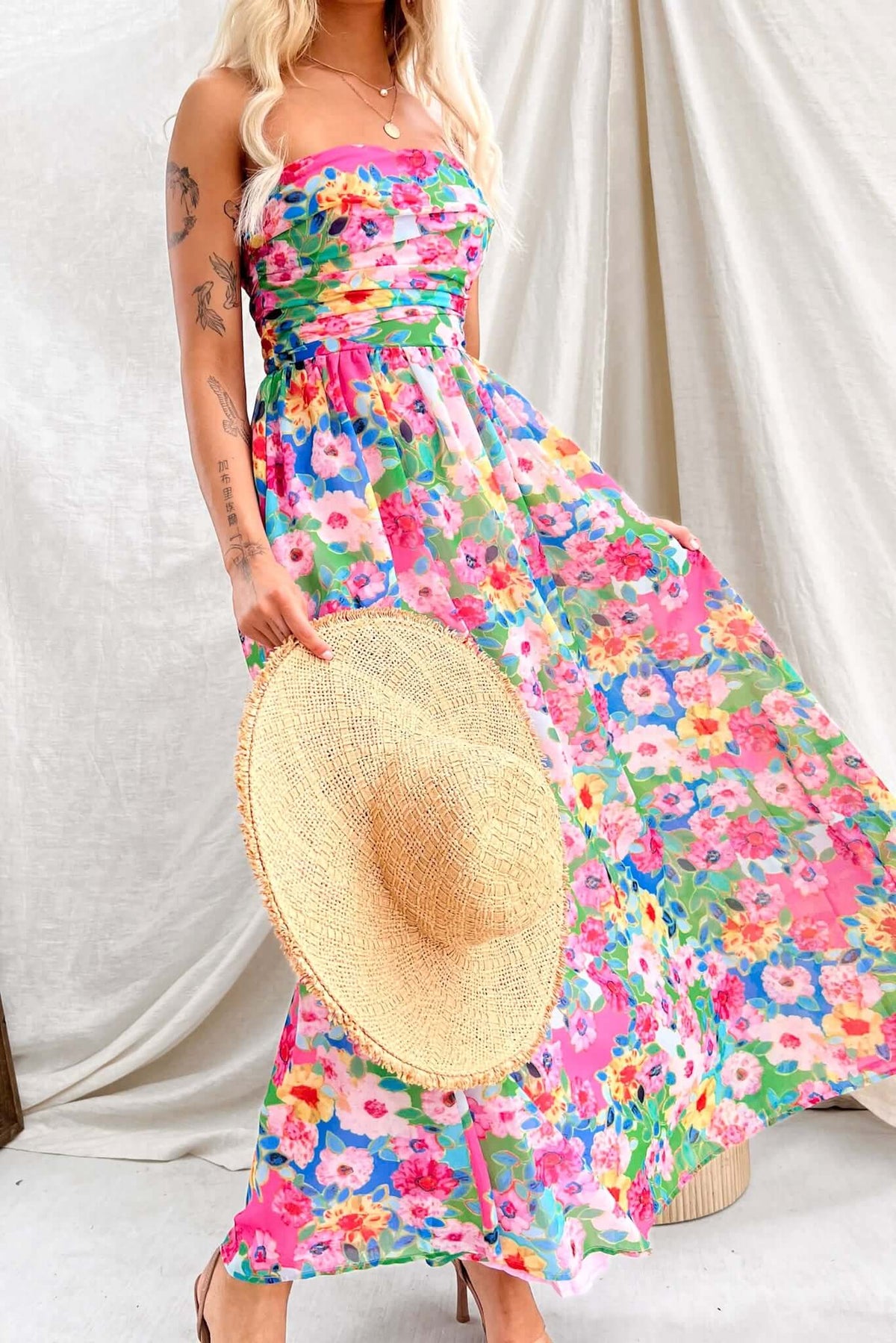 Model in bright floral Emberia Maxi Dress holding a straw hat, showcasing a strapless and flowing design.