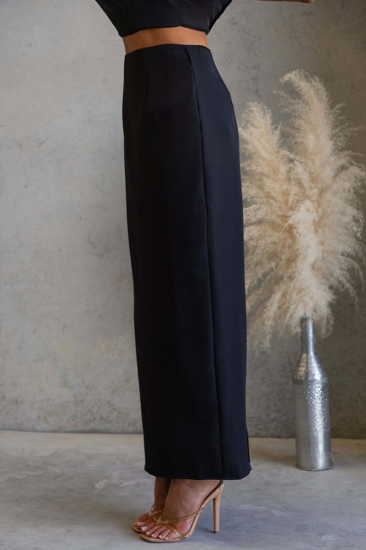 Duchess Maxi Skirt displayed on model, featuring black fabric and mid-high waist fit, styled with clear heels.