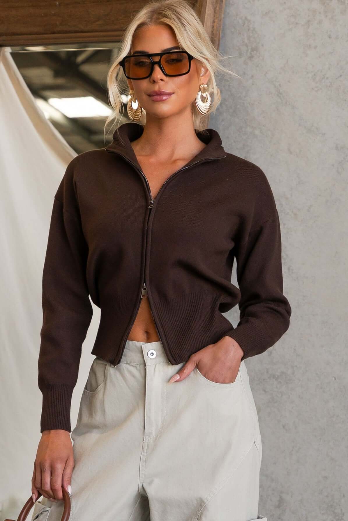 Model wearing a brown Grand Top with double zip-up front and mock neckline, styled with light trousers.