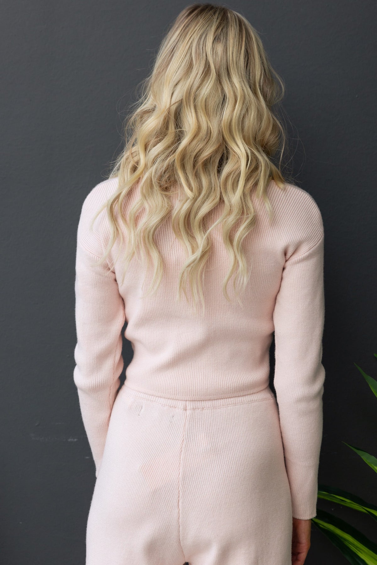 Back view of a woman in a pale pink ribbed knit top with long sleeves, styled with wavy blonde hair.