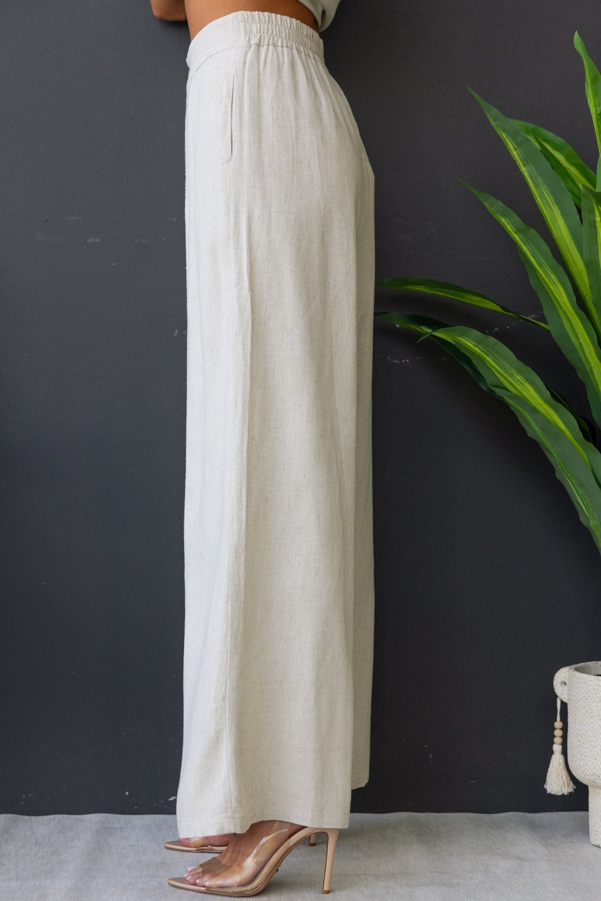 Bexley Pants in linen blend, featuring mid-high waist, elastic waistband, and pockets, styled with heels and greenery.